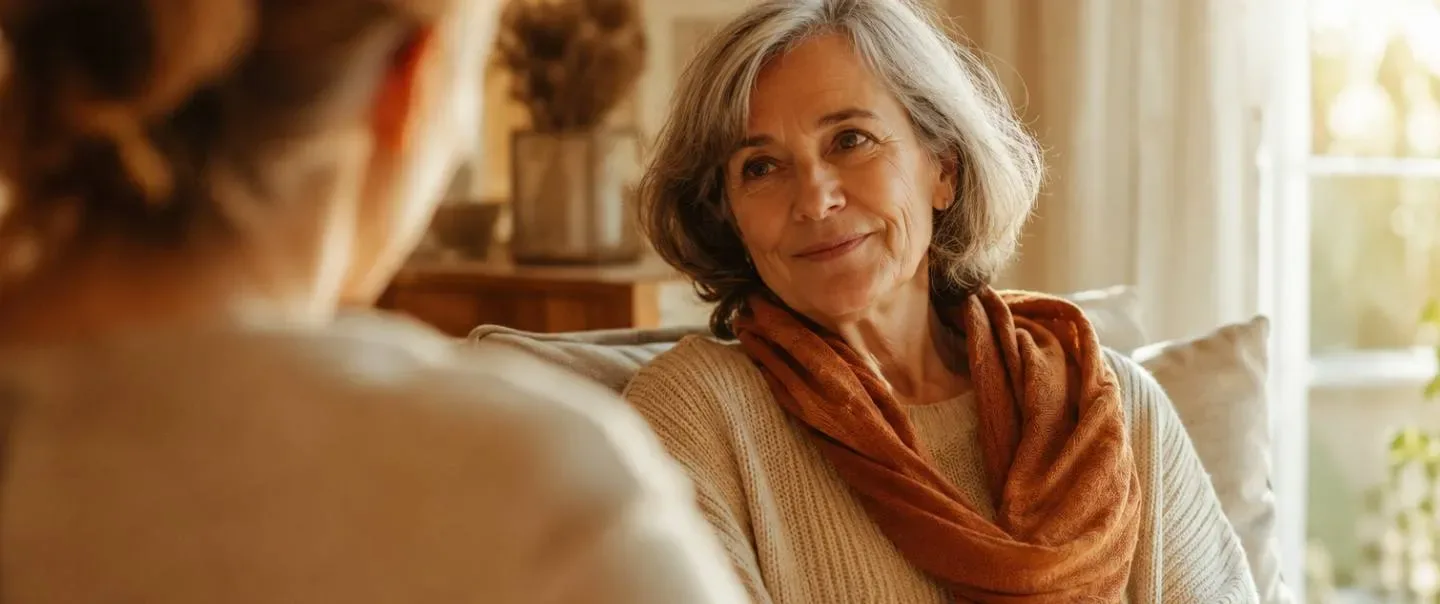 Dos mujeres conversan con calma en el salón de casa, una escucha a la otra con luz cálida de ventana